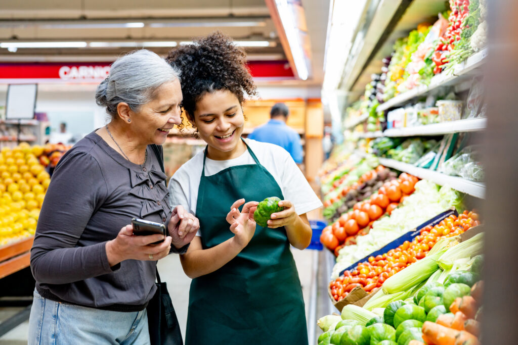 Friendly saleswoman showing a vegetable to senior woman who is holding a smart phone at the supermarket