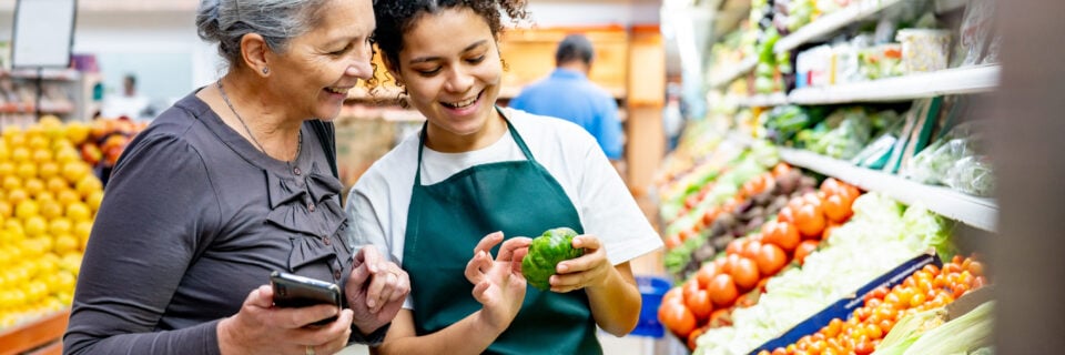 Friendly saleswoman showing a vegetable to senior woman who is holding a smart phone at the supermarket - Lifestyles concepts