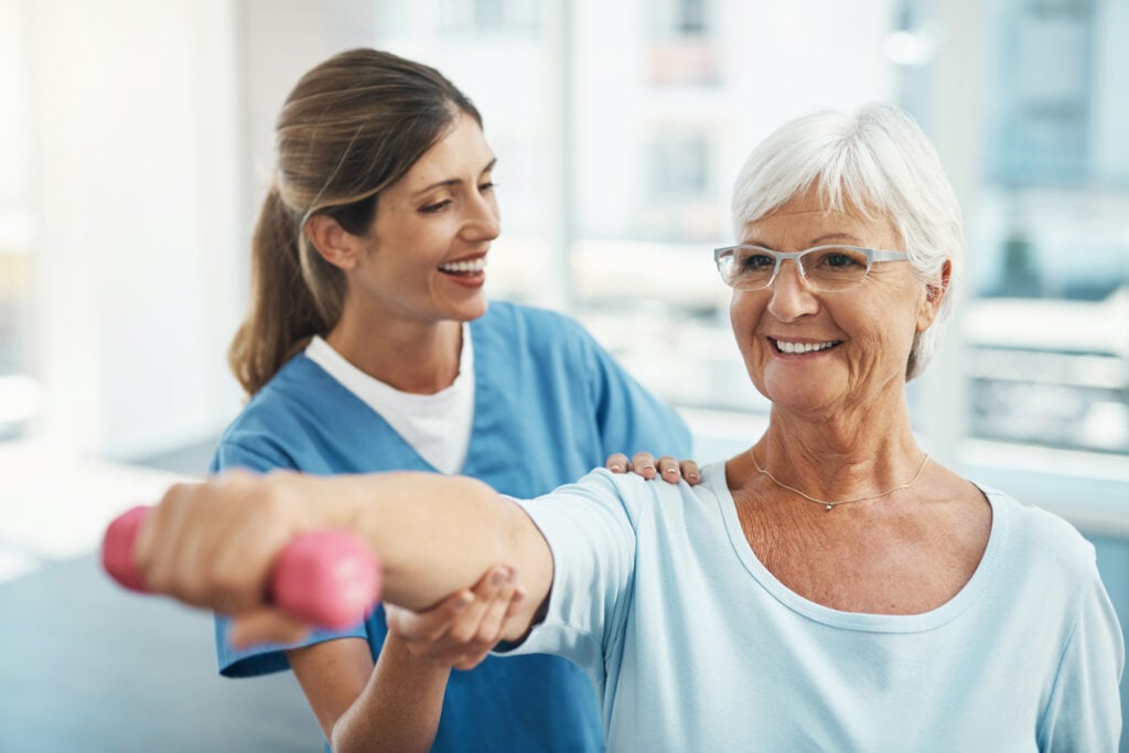 Senior woman with physical therapist lifting light pink dumbbell.
