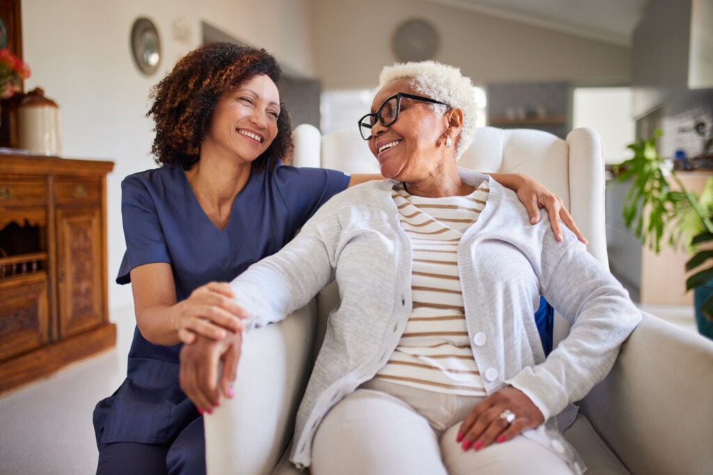 Senior woman and caregiver smiling at each other and sitting on an armchair.