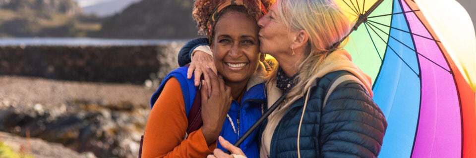 A waist-up shot of a same-sex female couple laughing under a multicoloured umbrella. The woman on the right is kissing her girlfriend on the side of the head. They are standing on the side of Loch Torridon near the village of Diabaig in the west highlands of Scotland.